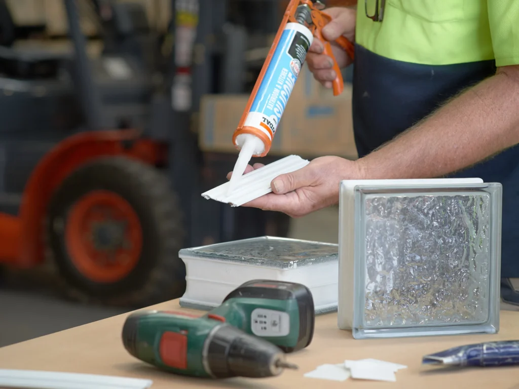 Worker applying sealant with a caulking gun to prepare the installation of glass bricks, with tools and materials on a construction workbench.