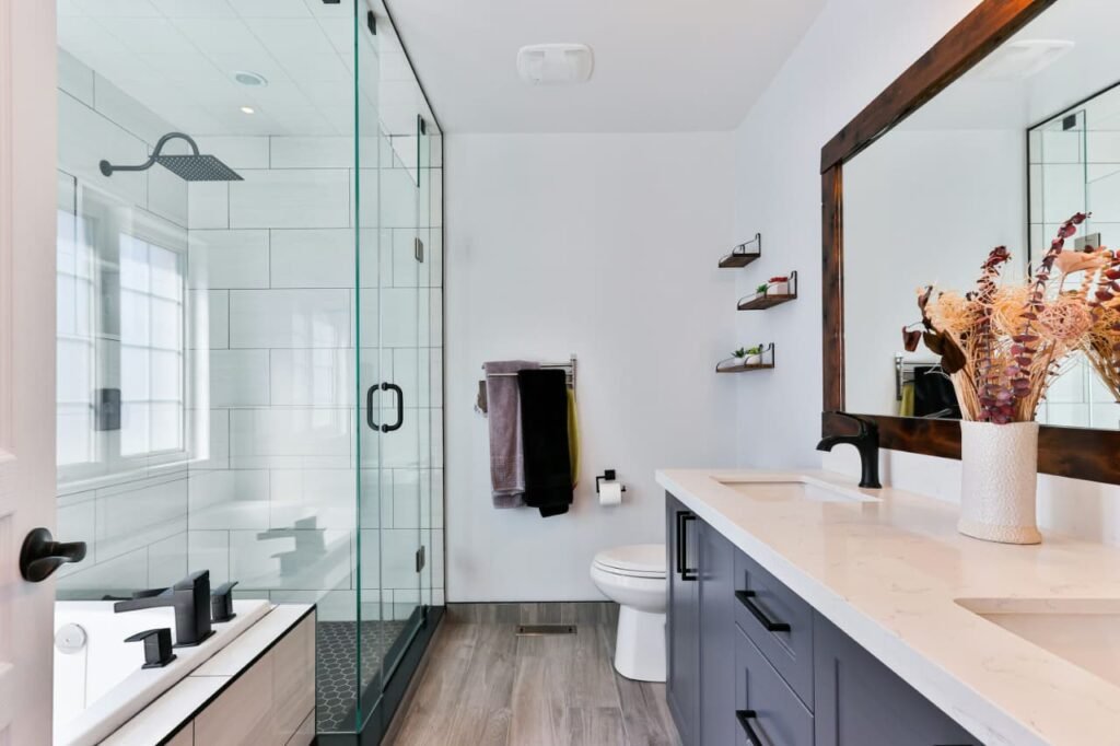 Modern small bathroom redesign ideas featuring a glass shower enclosure, wooden flooring, double vanity with white countertop, and stylish matte black fixtures for a contemporary, space-saving look.
