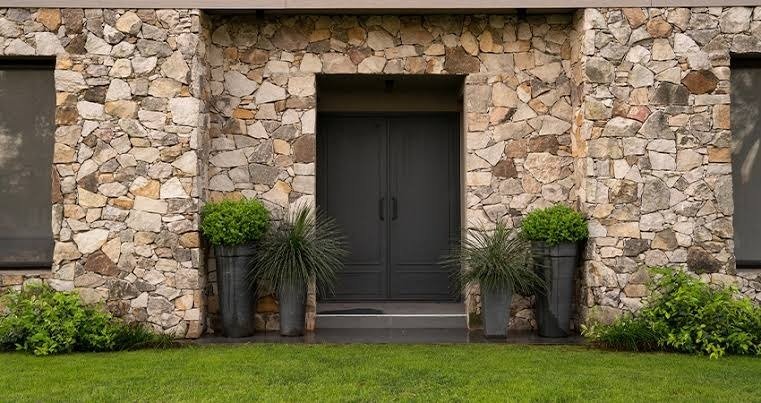 Front view of a luxury home in Gurgaon featuring designer exterior wall tiles in a natural stone look. The textured wall cladding surrounds a dark double door with potted plants, showing how outdoor tiles enhance a home’s entrance and add lasting style.