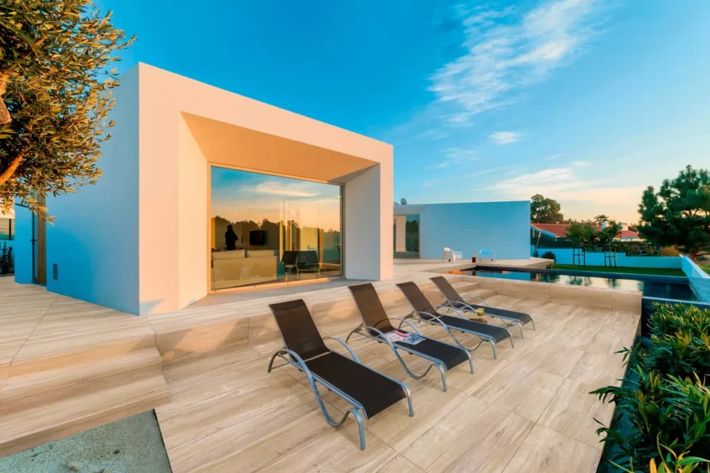 Modern villa exterior with light wood-look terrace tiles, poolside loungers, and a minimal architectural design at sunset.
