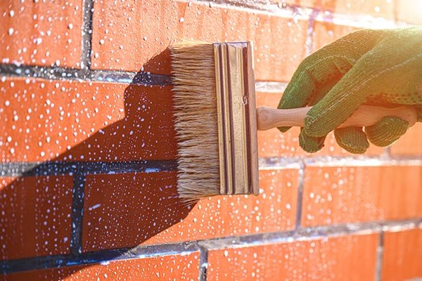 Close-up of vitrified tiles on an exterior brick-style wall being coated with a protective sealant using a wide brush, showing durability and low-porosity surface features of vitrified tiles.