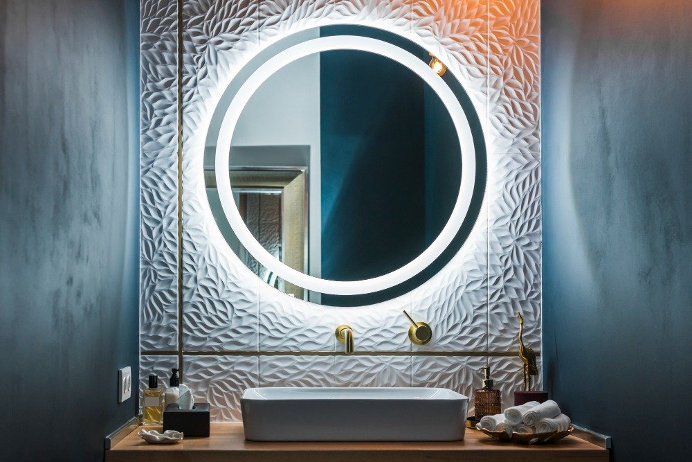 Elegant round LED mirror mounted on textured feature wall with premium basin and brass fittings, representing LED mirrors for modern bathrooms in upscale homes.